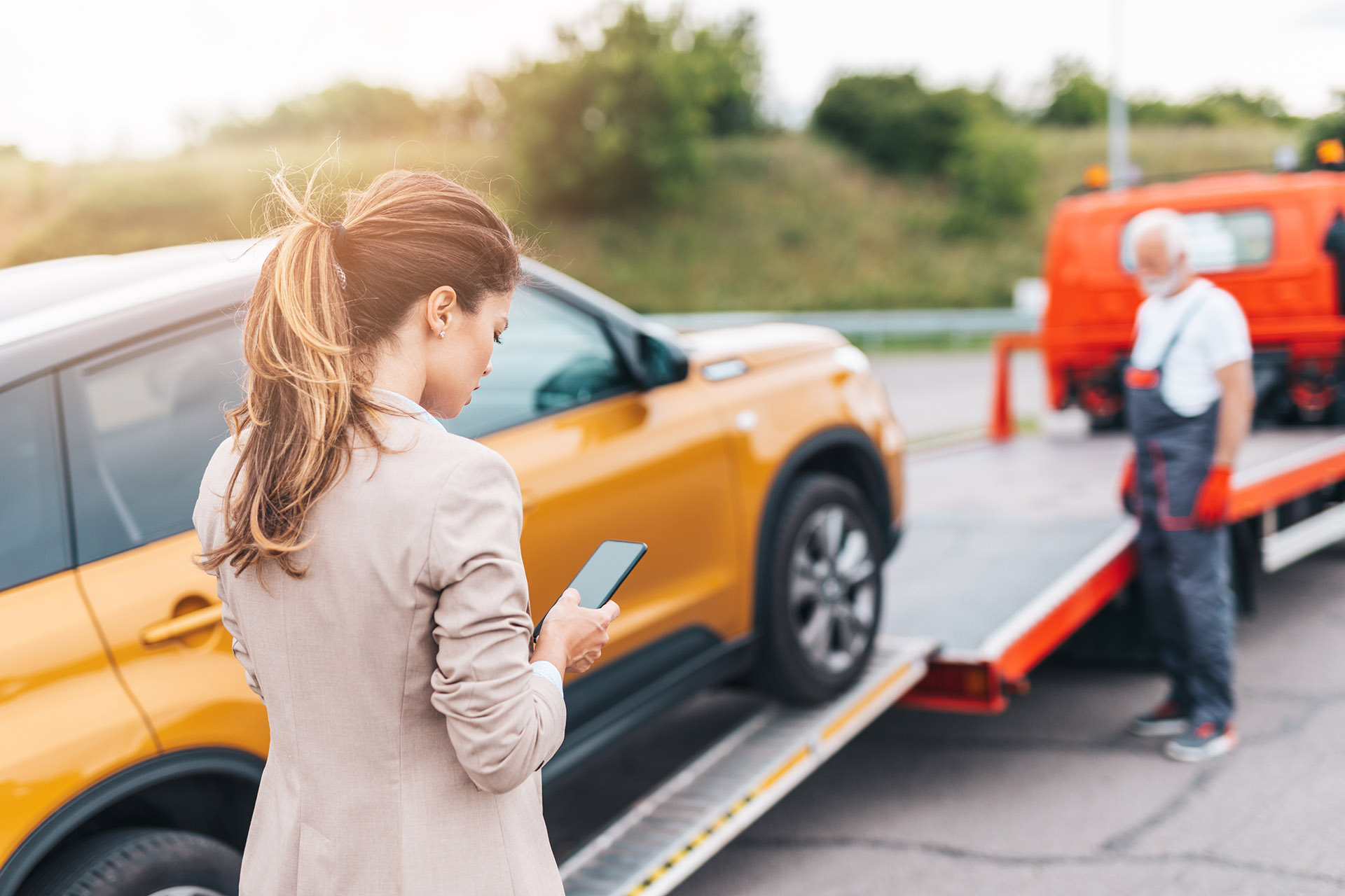 Elegant business woman making a phone call while towing service helps her on the road. Used to illustrate the article, "How Can a Denver Collision Center Help After a Car Accident."